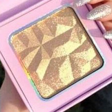 A close-up of the Nayy Seduction Brand's Girl Glow Highlighter in a geometric-patterned gold finish, housed in a pink square compact. This vegan and cruelty-free highlighter offers a mix of matte and shimmer finishes, resulting in a textured, faceted appearance. A person's manicured hand with glittery nails is holding the compact.