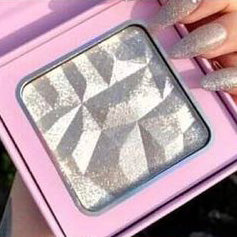 A close-up of the Girl Glow Highlighter by Nayy Seduction Brand, a compact and vegan makeup highlighter with a silver, faceted, gem-like design. The highlighter is housed in a pink case. A hand with glittering, silver-colored nails holds the case, showcasing the elegance of this cruelty-free product.
